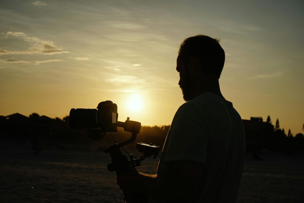 Videographer with camera equipment silhouetted against a sunset sky, capturing a serene outdoor scene.